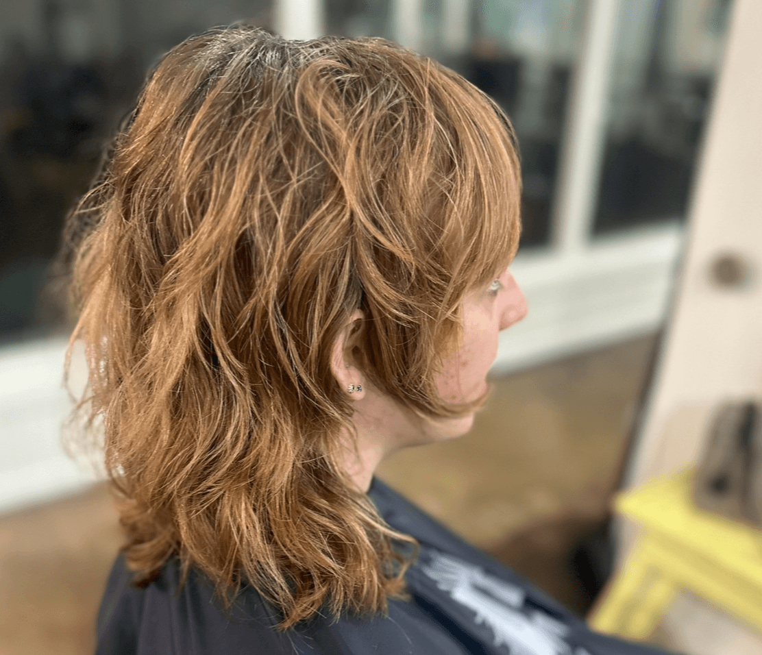 Side view of a person with wavy, shoulder-length hair, sitting indoors near a window.