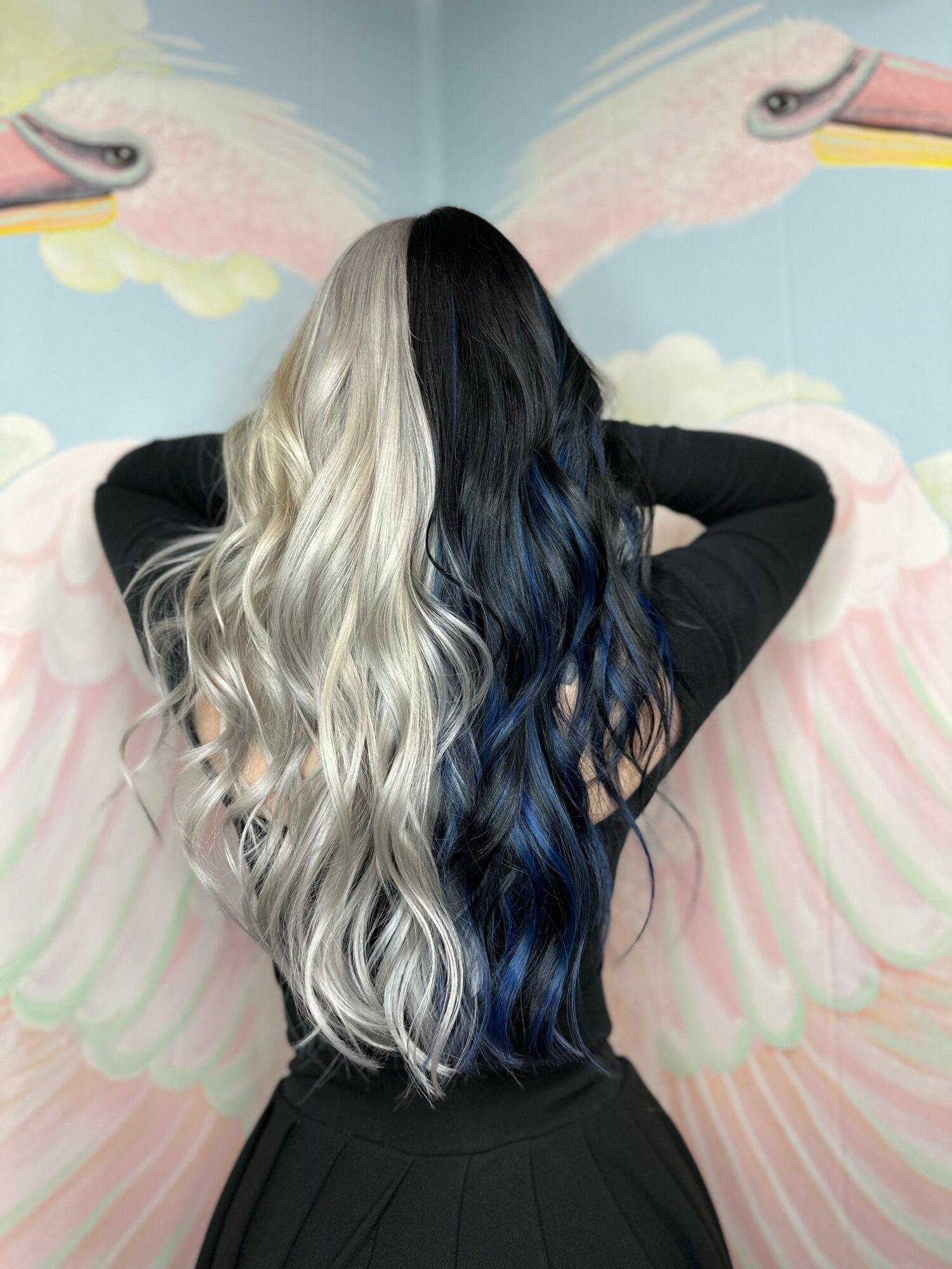 Person with long, wavy hair, half platinum blonde and half dark blue, standing with back to colorful swan mural.