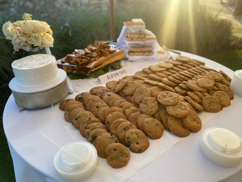 Table with gluten-free cookies, cakes, and snacks, decorated with flowers and sunlight, on a white tablecloth.