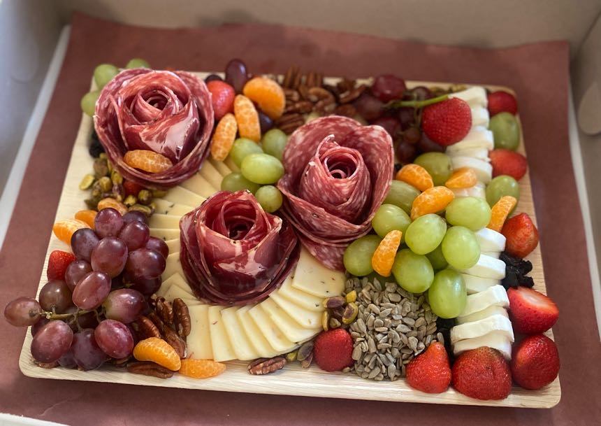 Decorative fruit and chocolate arrangement with three rose-shaped chocolates, grapes, strawberries, and assorted toppings on a tray.