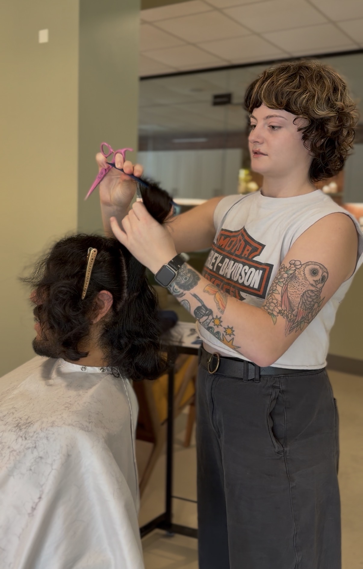 A hairstylist with tattoos and short curly hair styles a young girl's hair in a salon, using a comb and spray bottle.