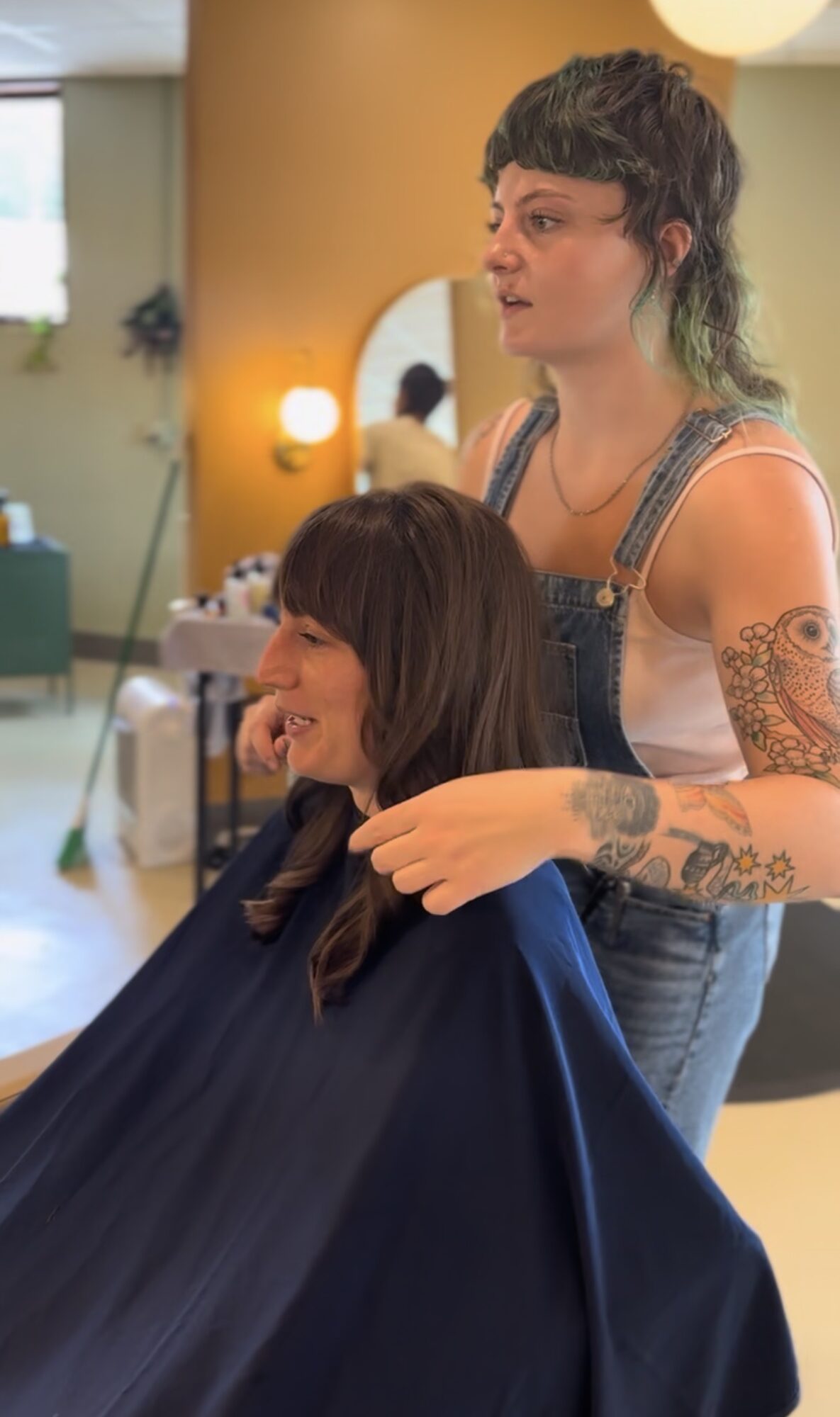 A hairstylist with tattoos and curly hair styles a woman with long dark hair in a salon. Bright, colorful background.