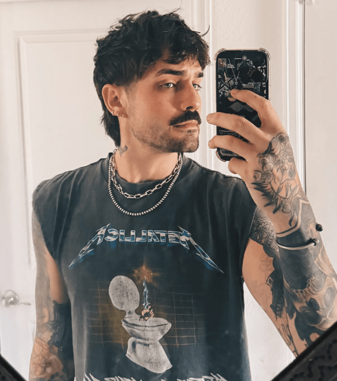 Young man with dark hair and tattoos taking a mirror selfie, wearing a sleeveless graphic T-shirt and layered necklaces.