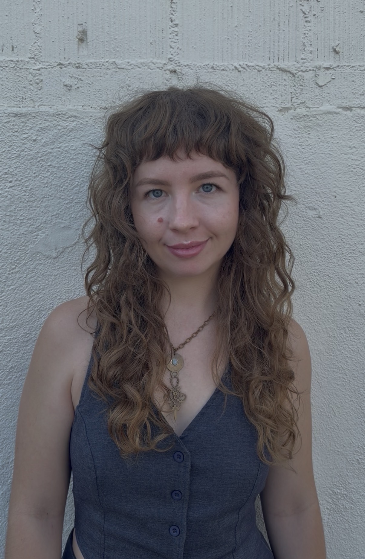 Young woman with curly hair and blue eyes, wearing a sleeveless dark top, standing against a light gray wall.