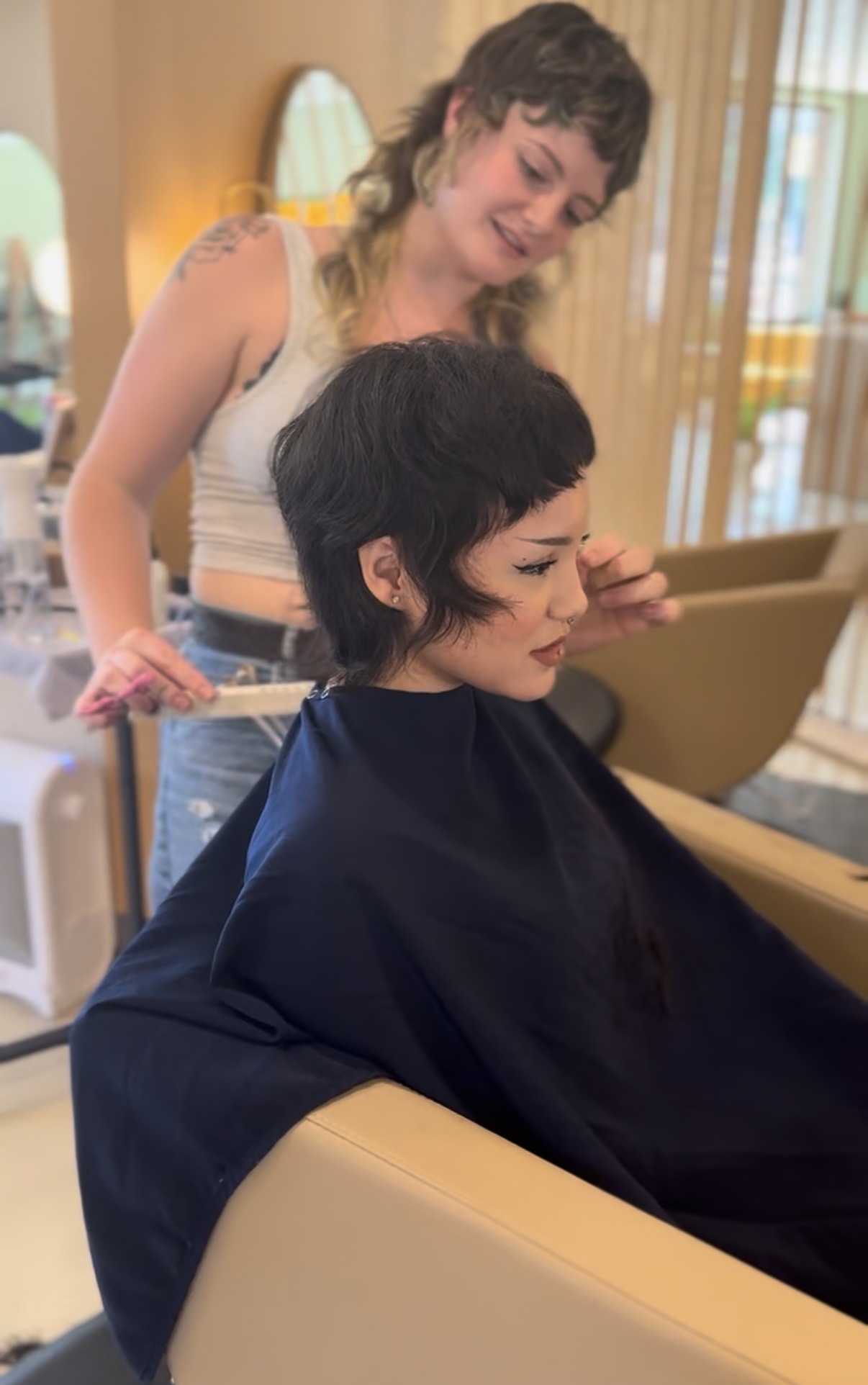 A woman with short black hair is seated in a salon chair, getting her hair styled by a stylist with curly hair.
