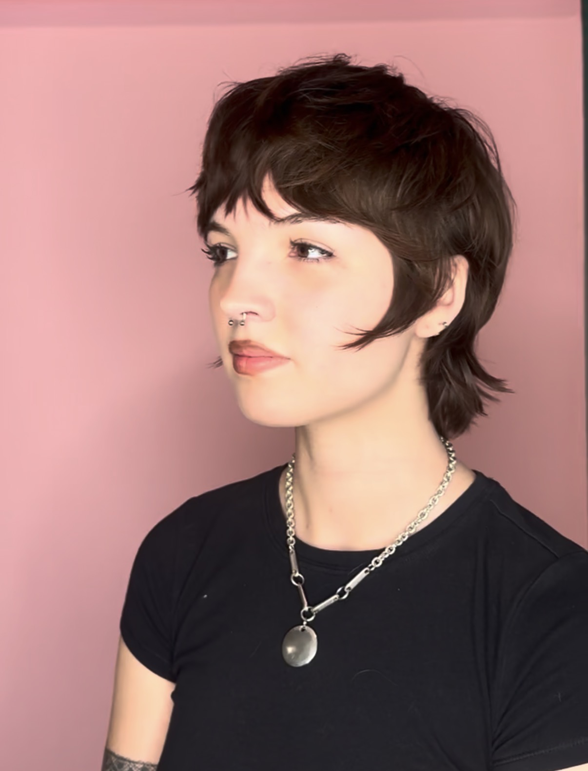 Young woman with short dark hair, wearing a black shirt and a silver necklace, looking to the side against a pink background.