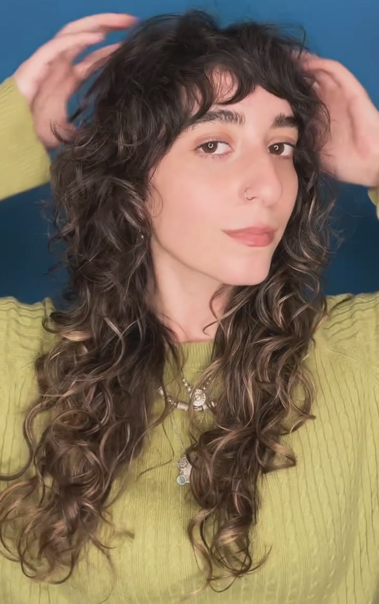 Young woman with curly hair and bangs, wearing a green sweater, posing with hands near her head, against a blue background.