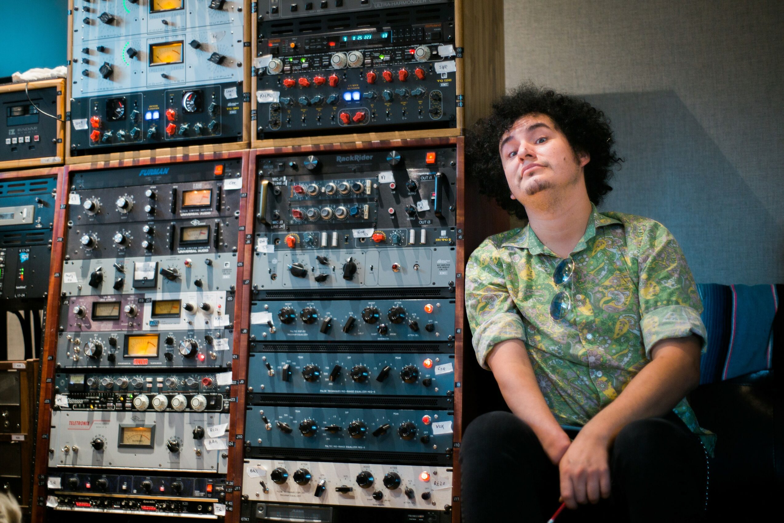 Person with curly hair sitting next to audio equipment racks, wearing a patterned shirt, looking at the camera.