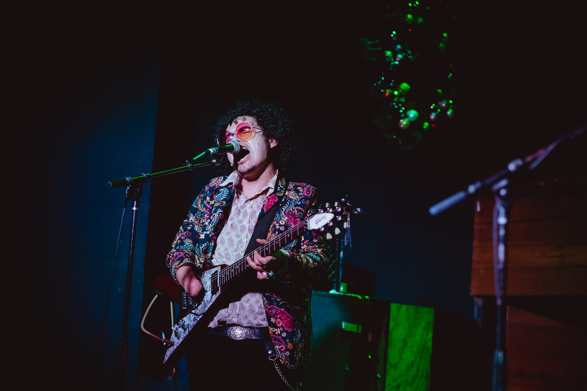 Person with curly hair and glasses playing guitar on stage, wearing a patterned jacket, with a microphone and green lighting in background.