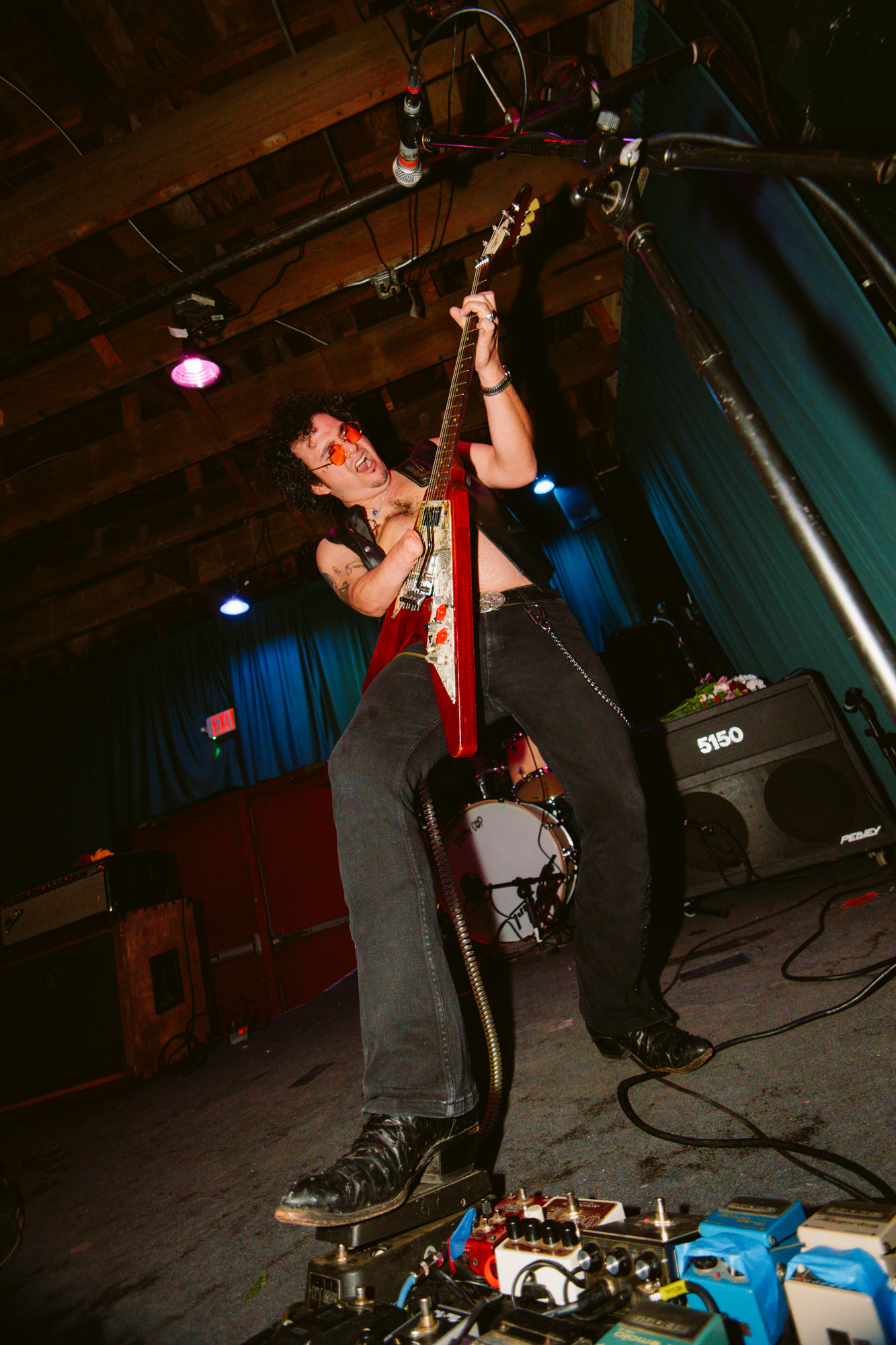 Person playing guitar on stage with equipment and colorful lighting, standing with one foot on a pedalboard, in a performance setting.