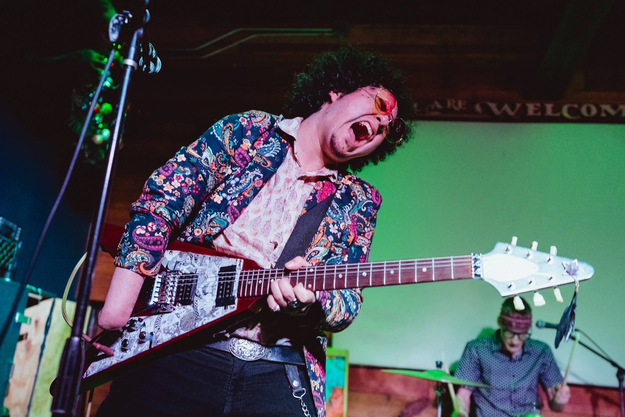 Person playing an electric guitar on stage, singing passionately with a colorful jacket, in front of a green screen background.
