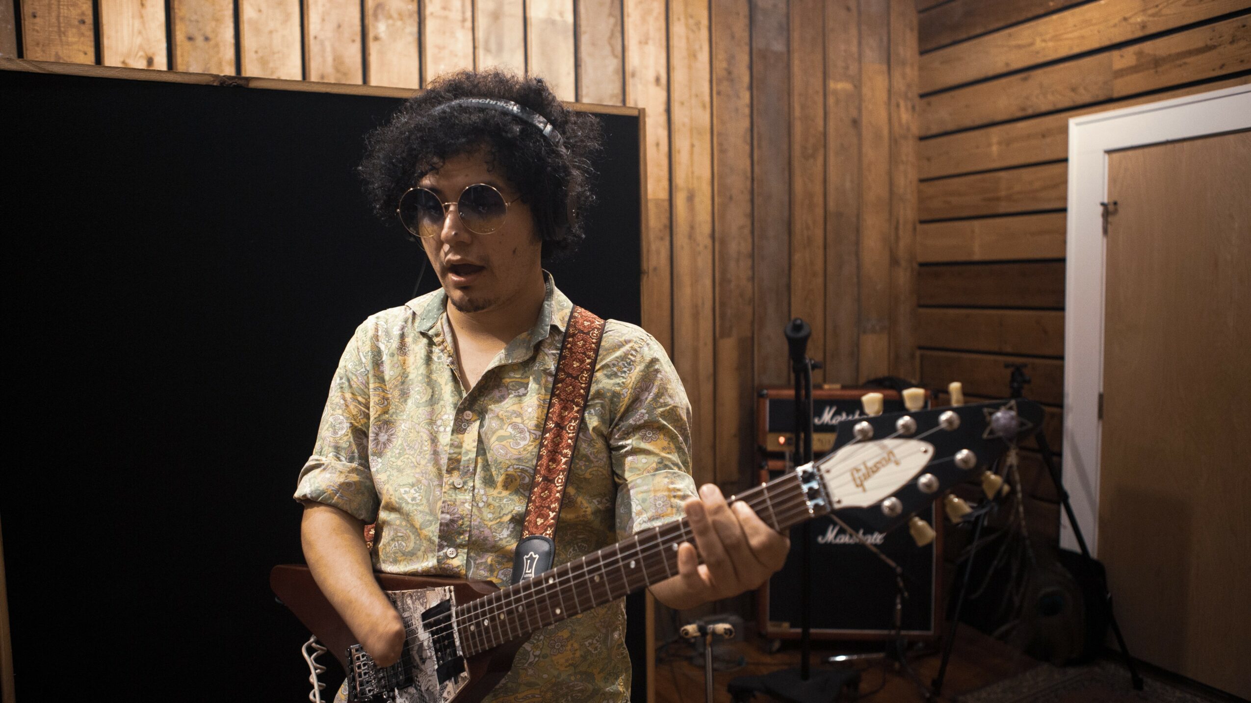 Person with curly hair wearing sunglasses and patterned shirt playing an electric guitar in a wooden room.