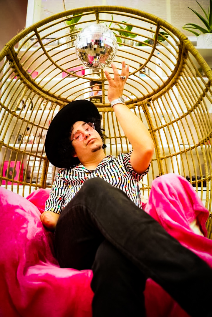 Woman with black hat and striped shirt reclining on pink fabric, reaching towards a reflective disco ball above her.