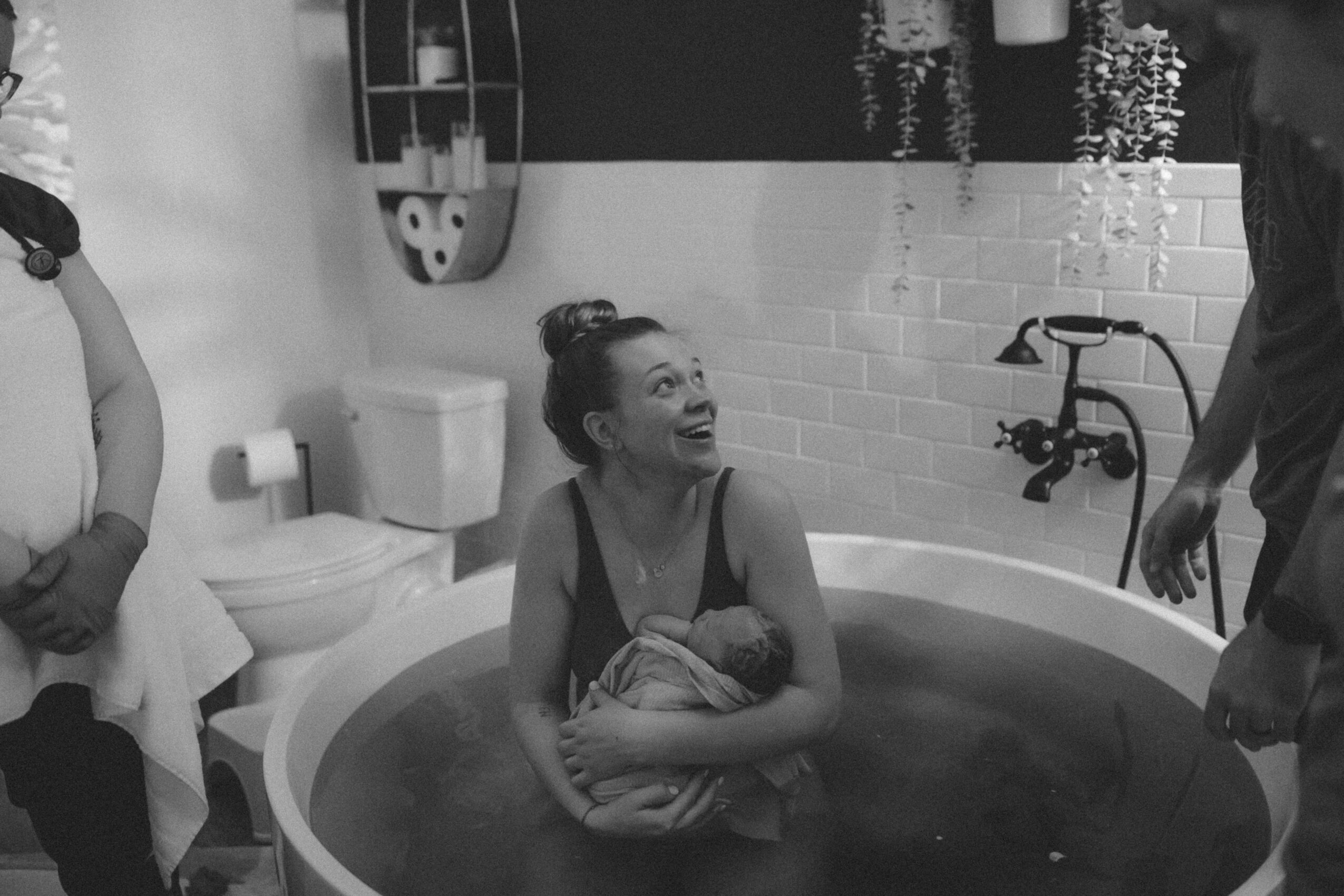 Woman sitting in a bathtub holding a towel, smiling, with two people nearby in a bathroom, black and white photo.