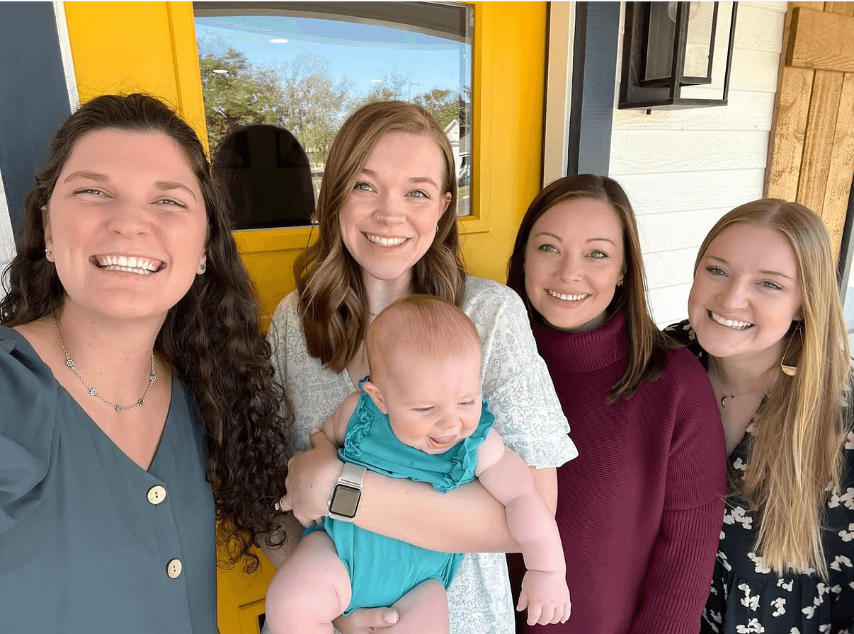 Four women and a baby smiling outside a building with a yellow door, one woman holding the baby.