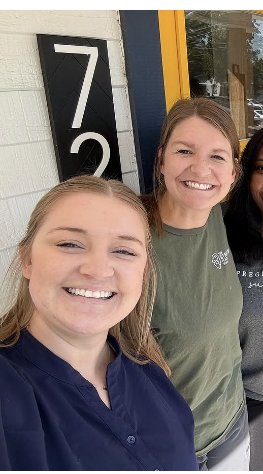 Two smiling women taking a selfie outside a building with a yellow and black sign, one wearing a green shirt, the other in navy blue.