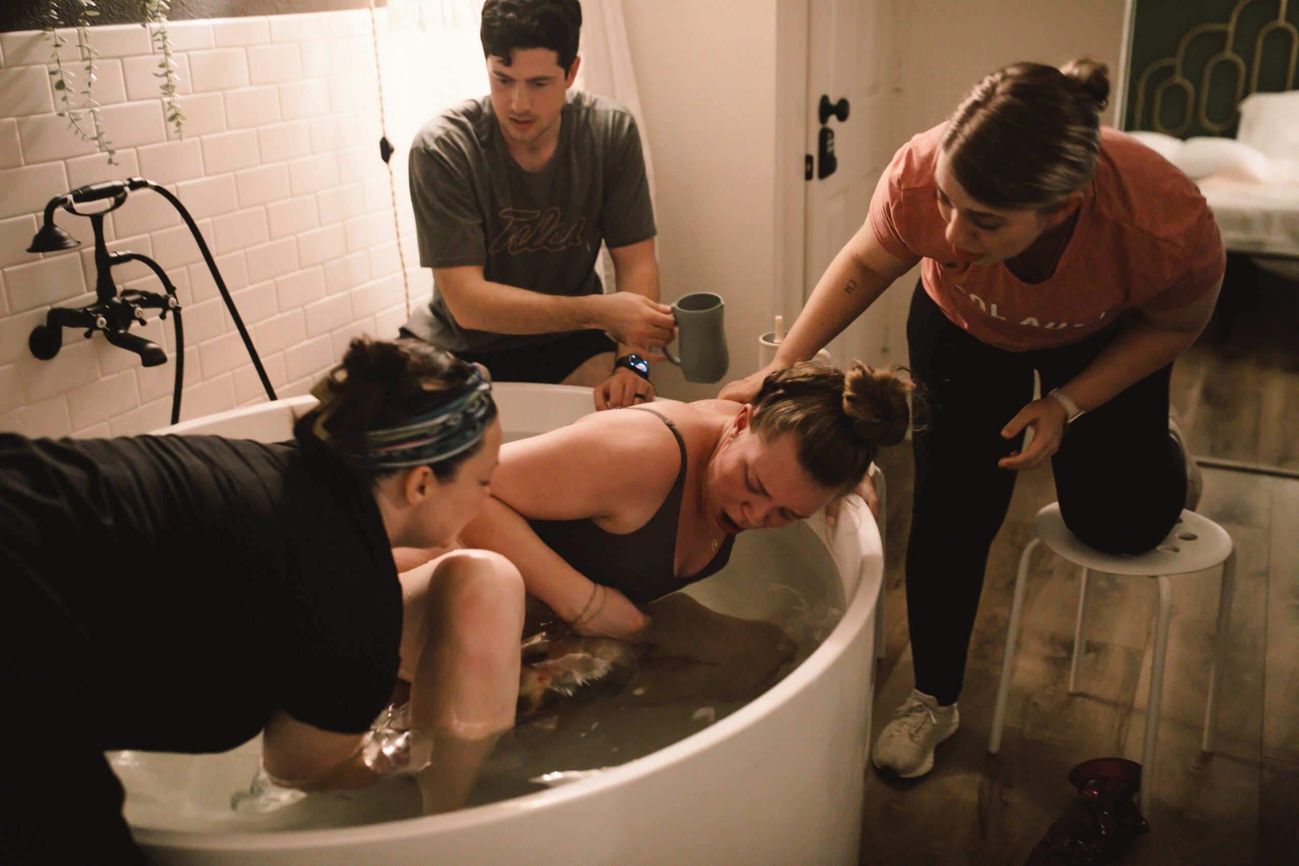 Four people in a bathroom, with one person in a bathtub, others assisting and observing, warm lighting, tiled wall background.