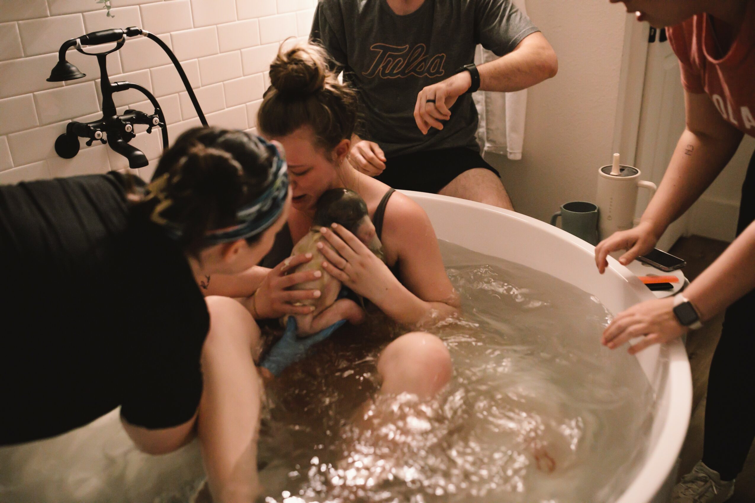 Person being baptized in a bathtub surrounded by four people assisting, with a showerhead visible, in a bathroom.