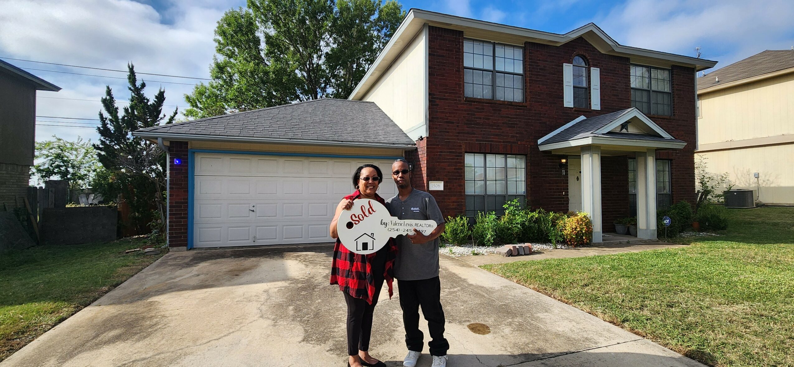 Two people stand in front of a two-story house holding a large key, with a driveway and lawn visible.
