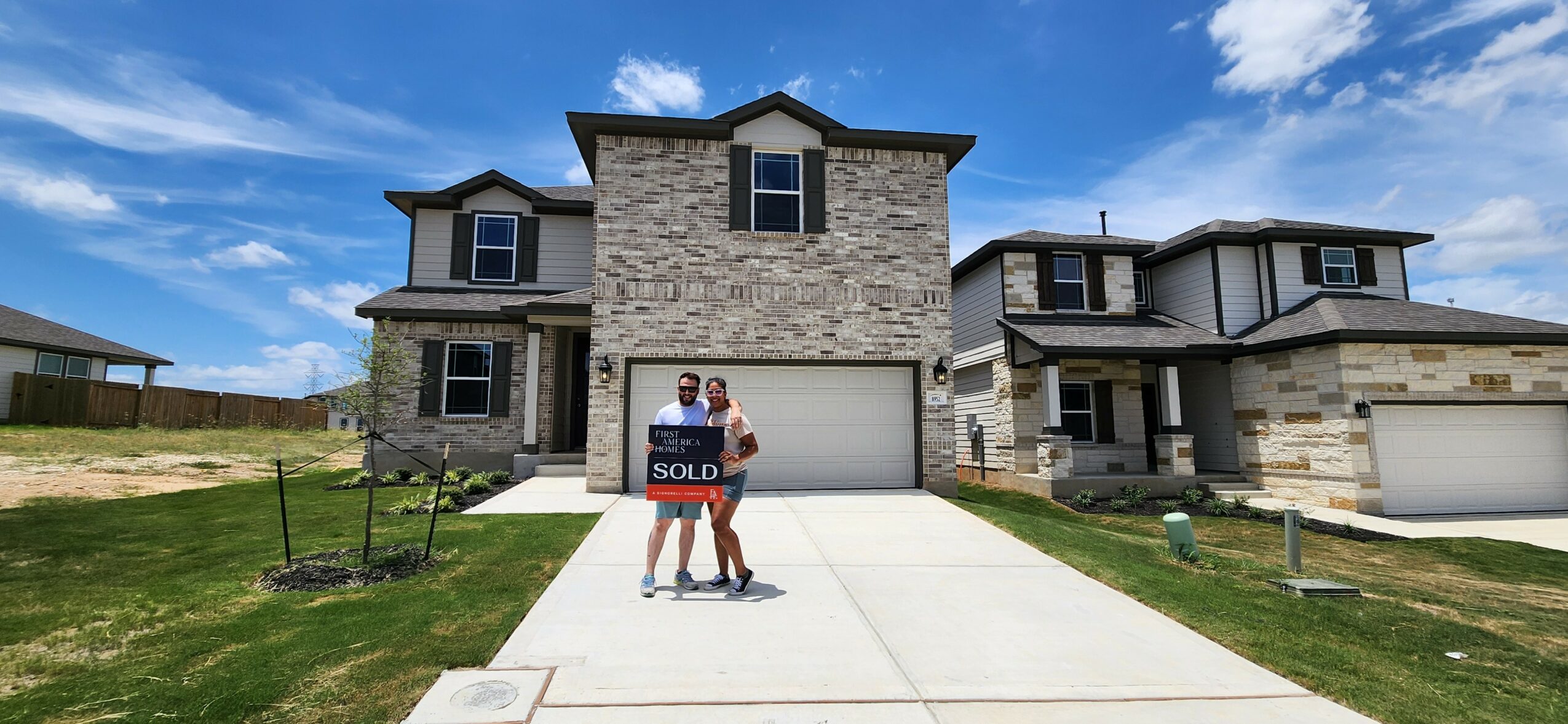 Two people holding a 'sold' sign in front of a new house with a driveway and neighboring homes, under a blue sky.