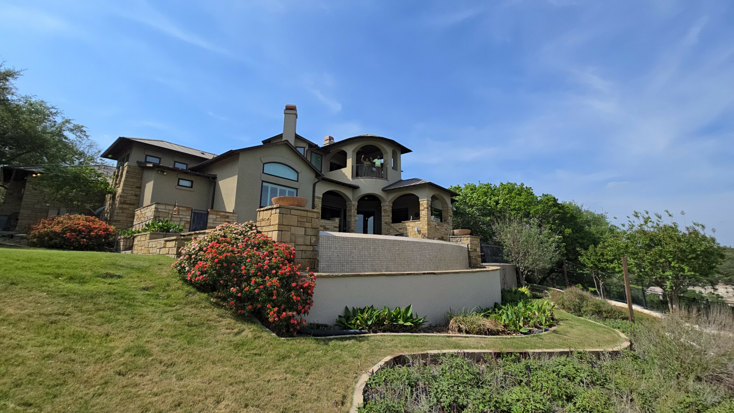 Large house with stone and stucco exterior, surrounded by greenery and flowering bushes, under a blue sky.