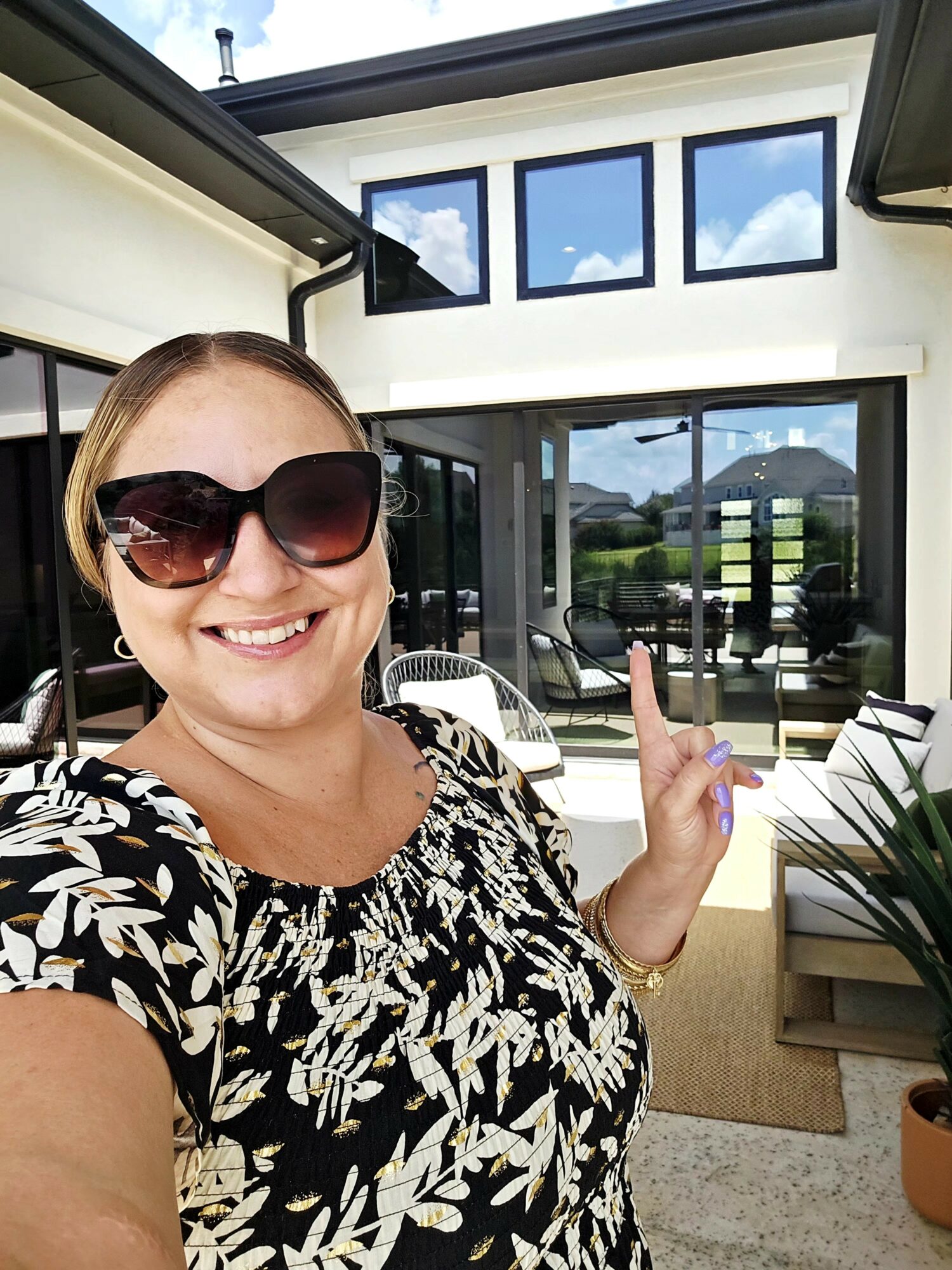 Woman with sunglasses smiling outdoors, pointing at a house with large windows and a patio.