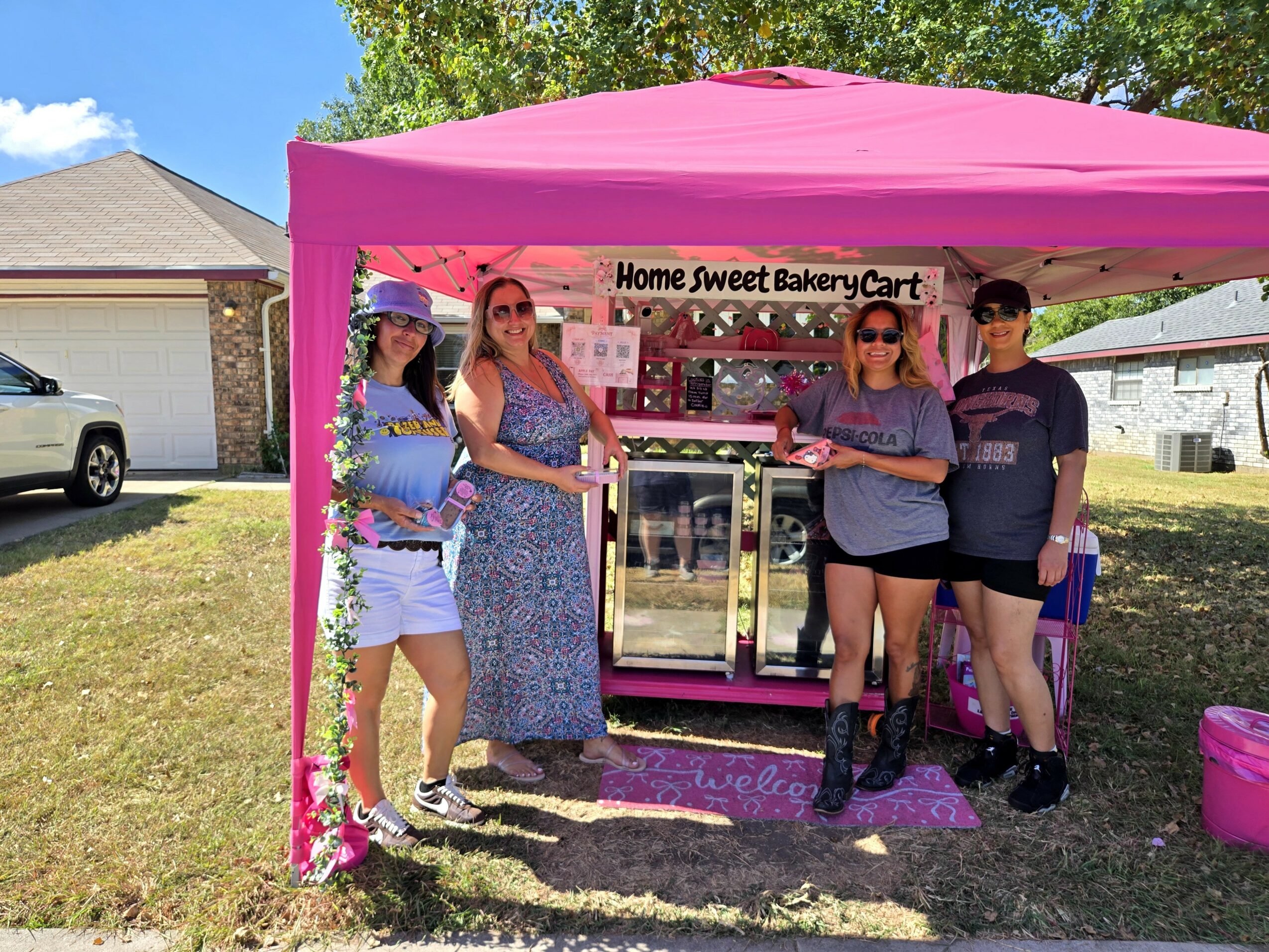 Four women stand in front of a pink tent with a bakery cart sign, outdoors on a sunny day.