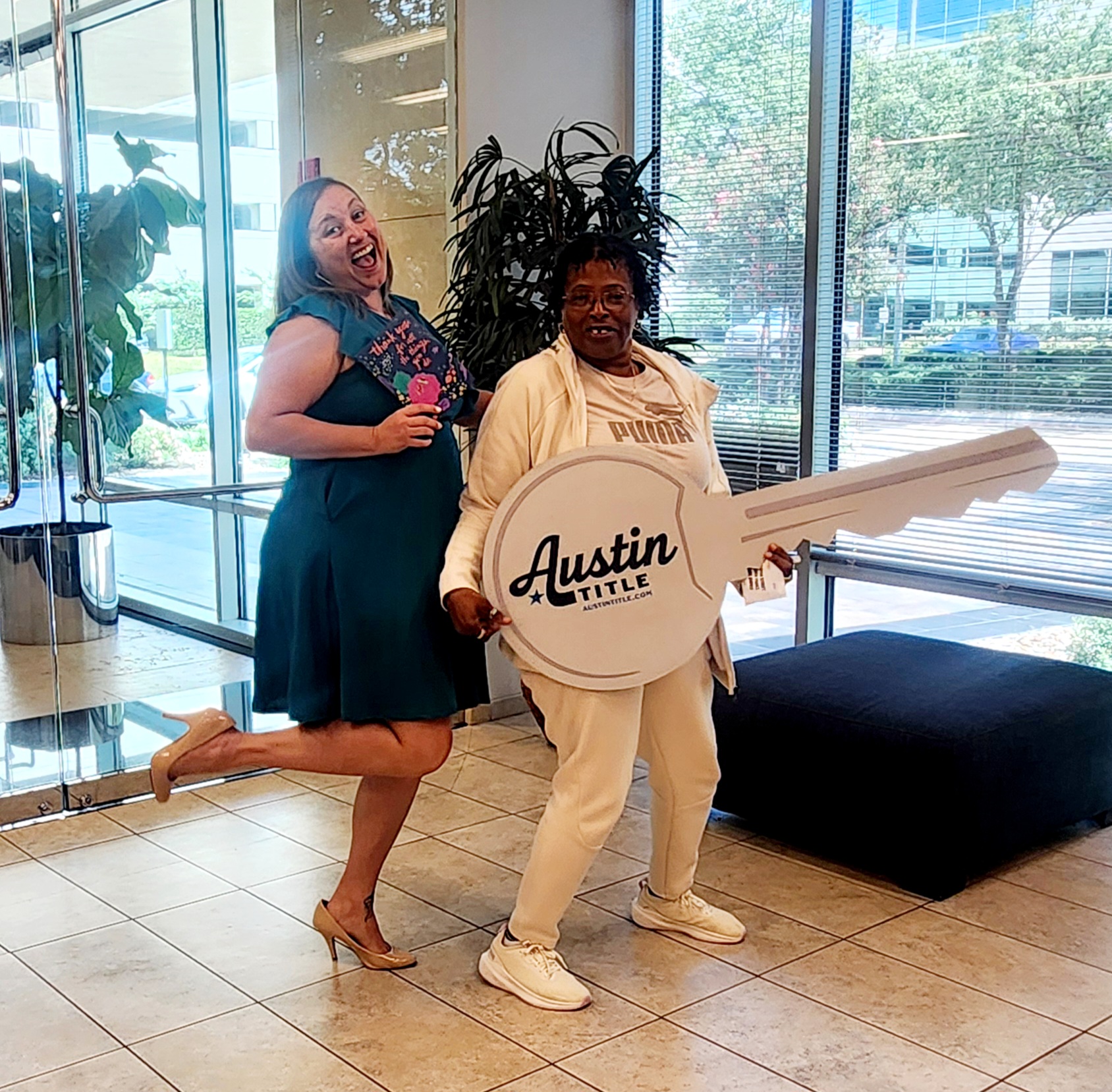 Two women posing indoors, one holding a large key with 'Austin TITLE' written on it, large windows in background.