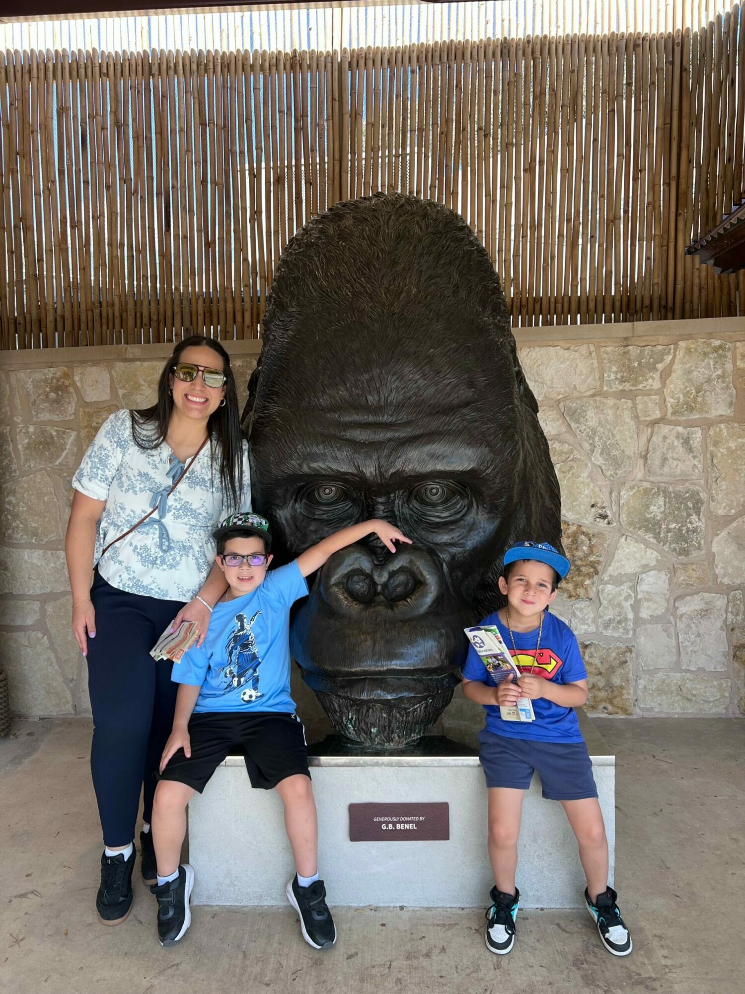 Two children and a woman pose with a large gorilla statue outdoors, with a stone wall and wooden fence behind them.