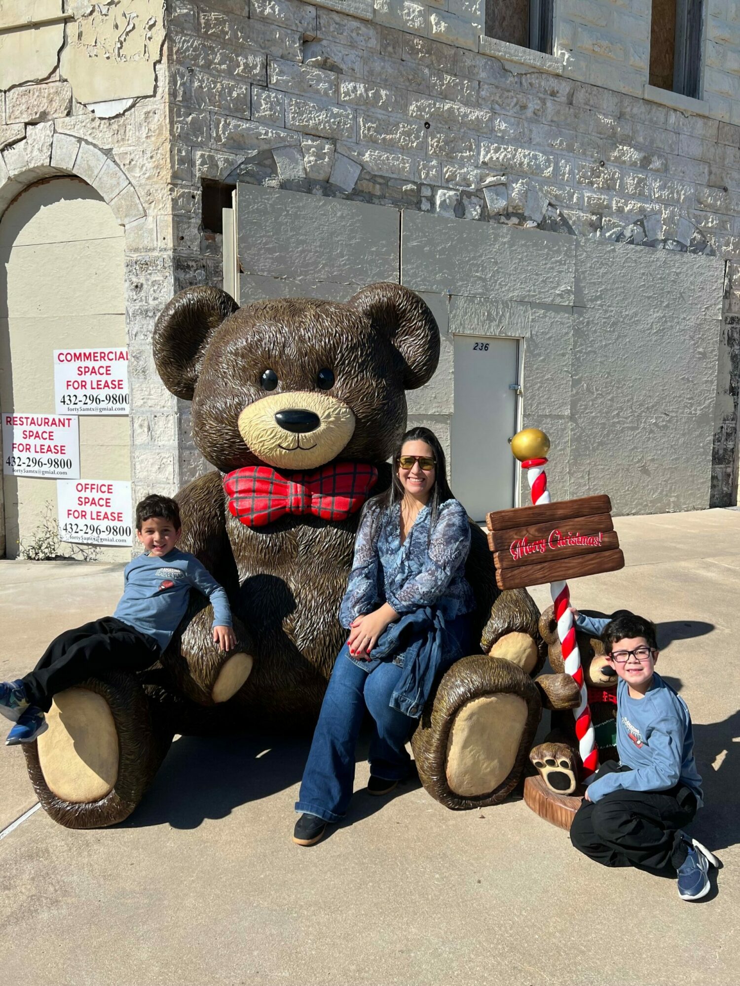 Person in a bear costume sitting on the ground with two children, one standing and one sitting, outdoors near a stone wall.