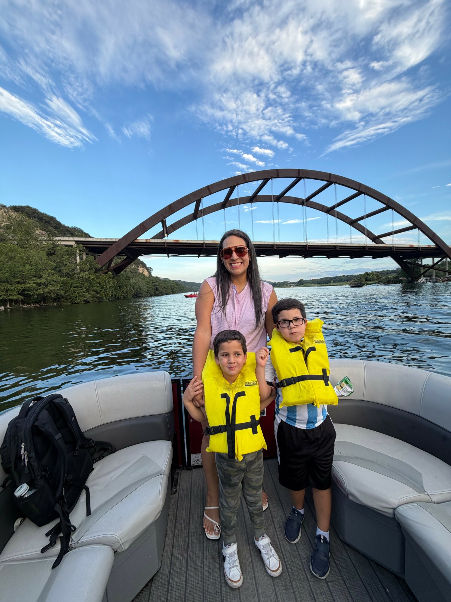 Woman and two children wearing yellow life jackets on boat with bridge and water in background.