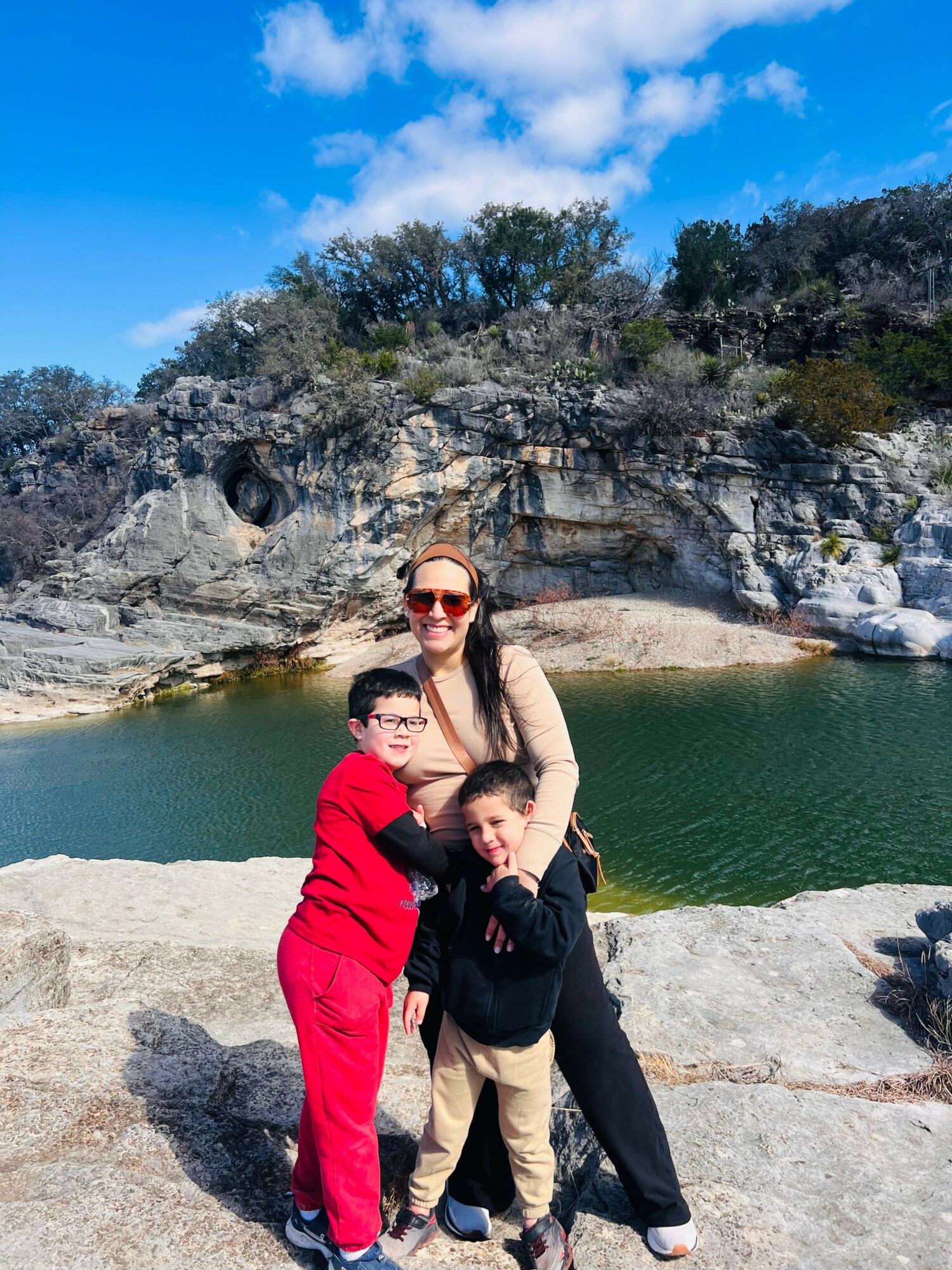 A woman with two children standing on a rocky shoreline near a body of water, with cliffs and trees in the background.