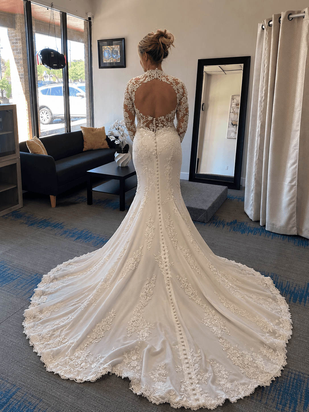Woman in a wedding dress with a long train, standing indoors near a mirror and window, facing away from the camera.