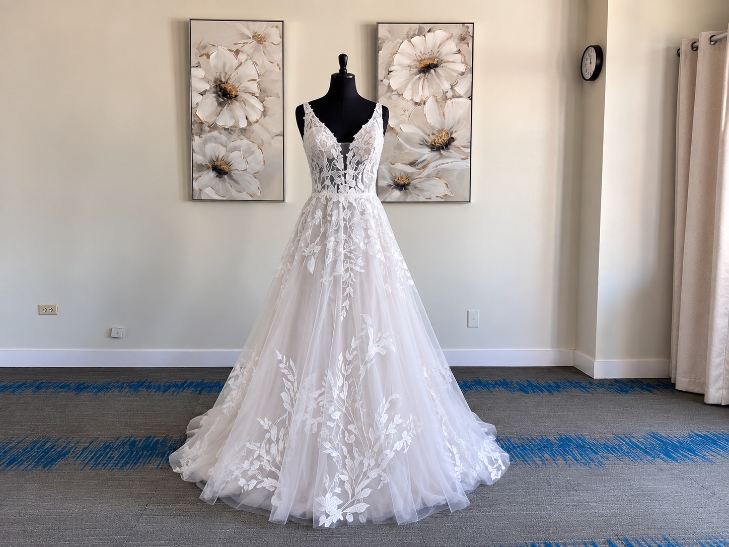 Wedding dress on mannequin with floral wall art behind, in a room with curtains and a clock, floor with blue stripe pattern.