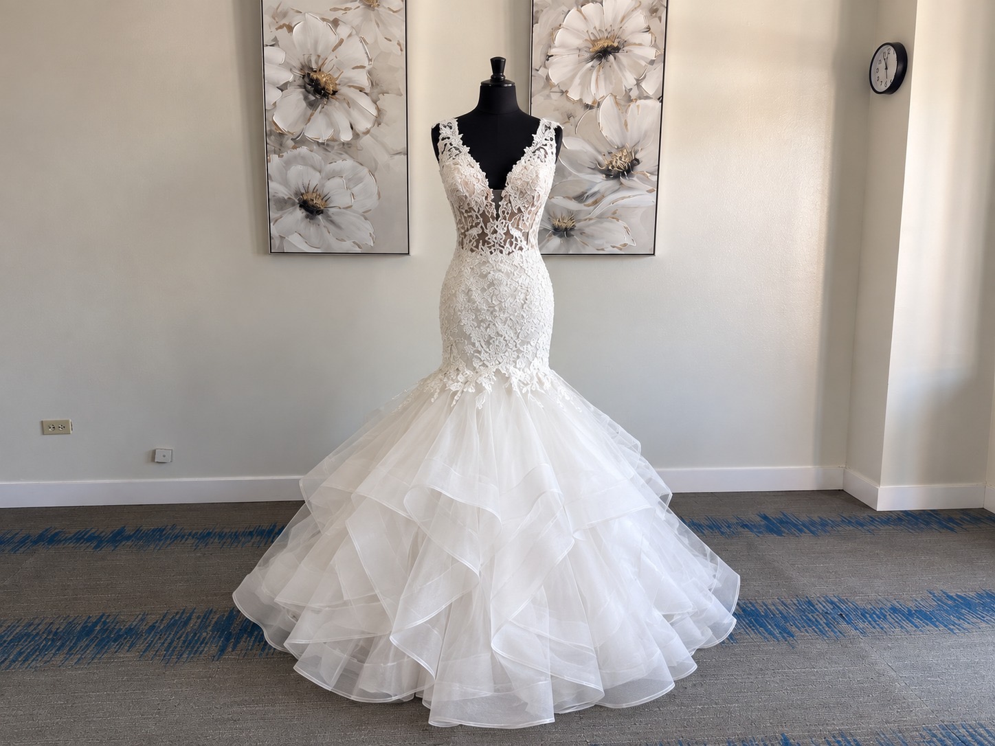 Wedding dress on mannequin with floral artwork behind, floor-length with layered tulle skirt, fitted bodice, and lace details.