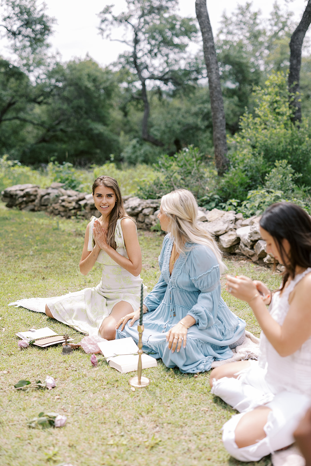 Three women sit on grass in a garden, engaging in a spiritual or meditative activity with candles and flowers.