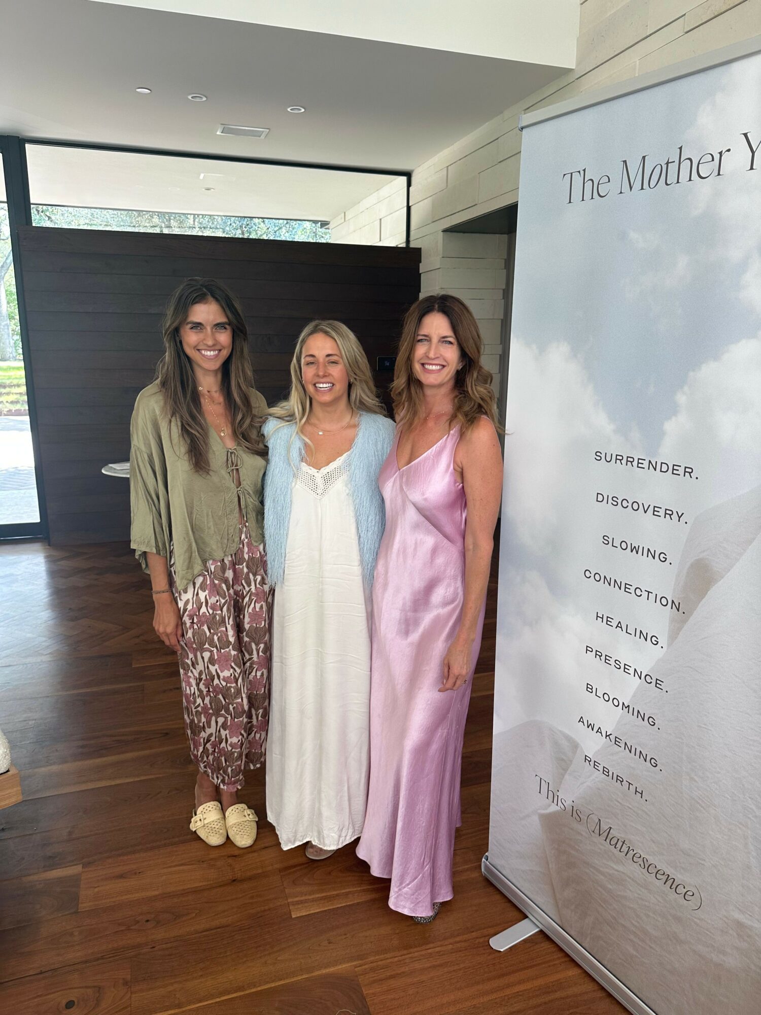 Three women standing indoors, smiling, with a large informational sign behind them. One woman wears a pink dress, another a white dress, and the third a patterned dress.