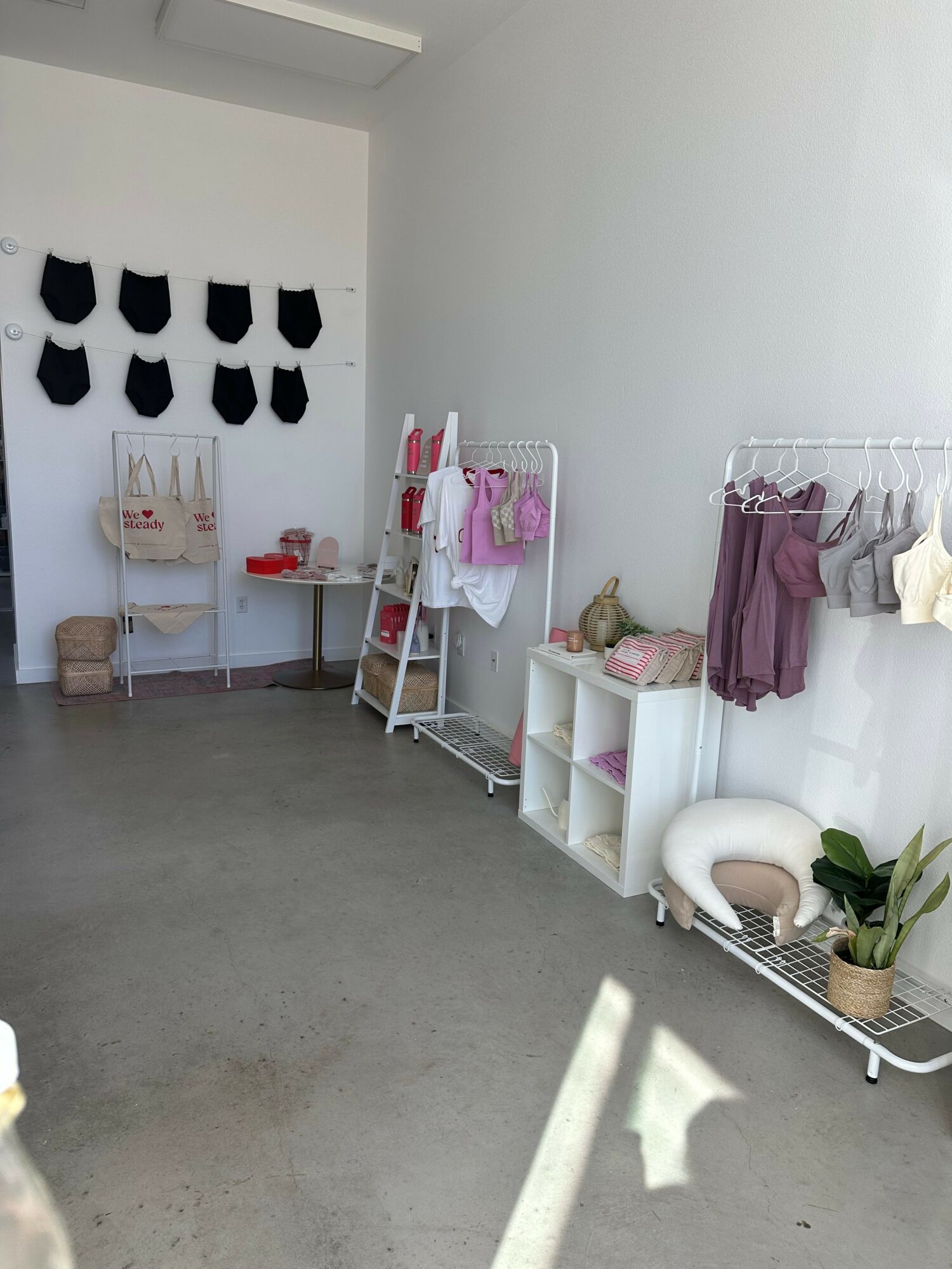 Display of clothing and accessories on white racks and shelves, with a potted plant and decorative items nearby.
