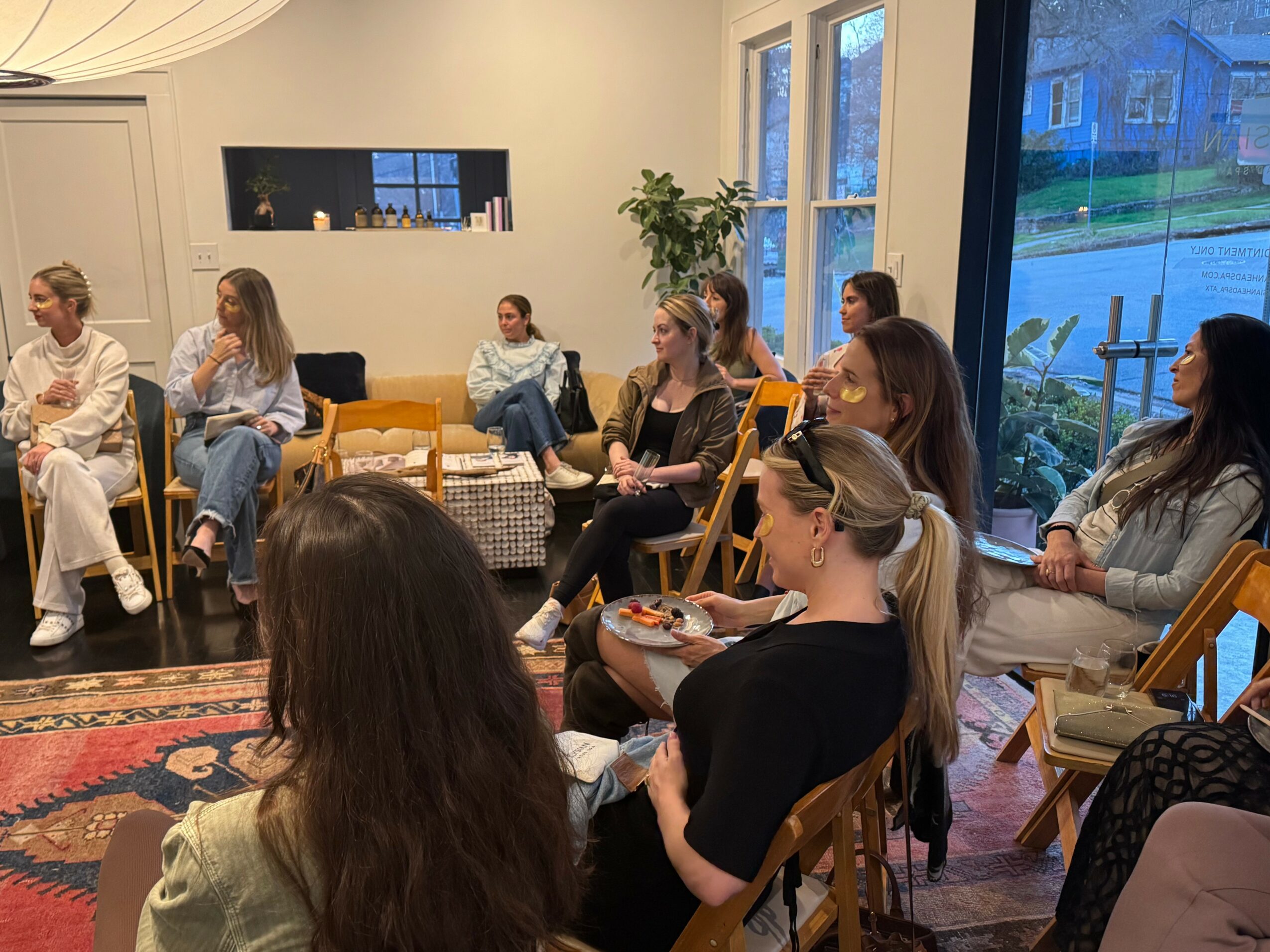 Group of women sitting in a living room, some holding plates, near large windows with a view outside.