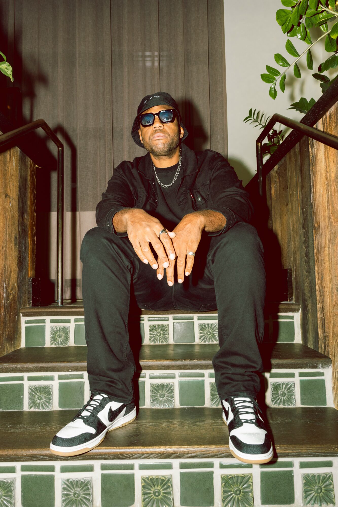 Man sitting on stairs with sunglasses, black jacket, black pants, and sneakers, hands resting on knees, indoors with plants nearby.