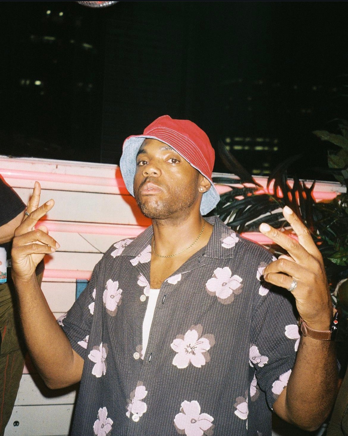 Man wearing a red bucket hat and floral shirt, making hand gestures, standing outdoors at night with city lights in background.