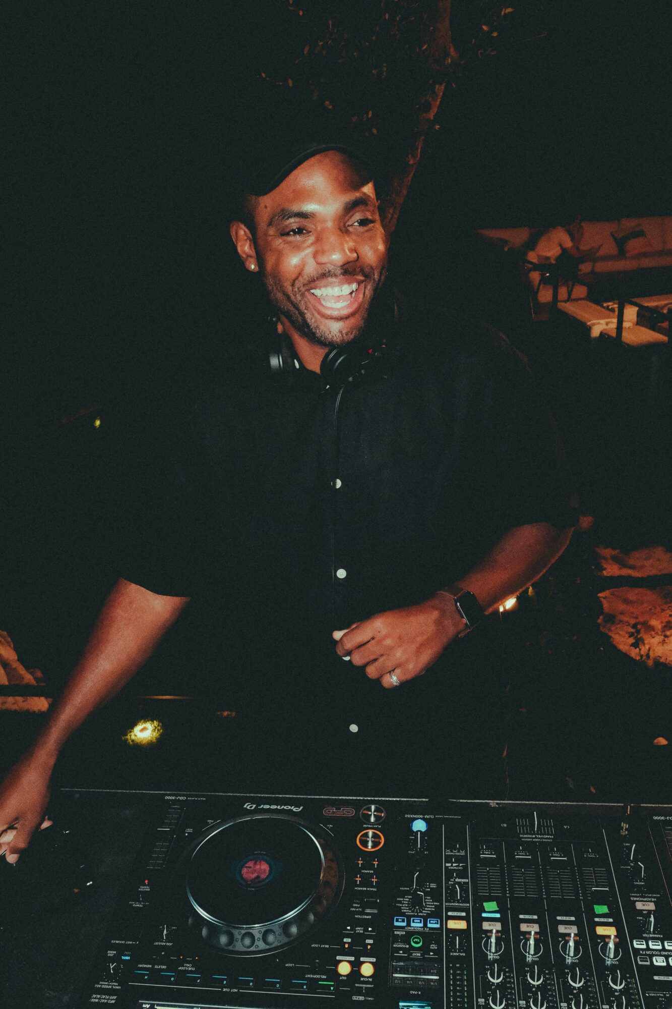 Man smiling, wearing a black shirt, standing behind DJ equipment in a dark setting.