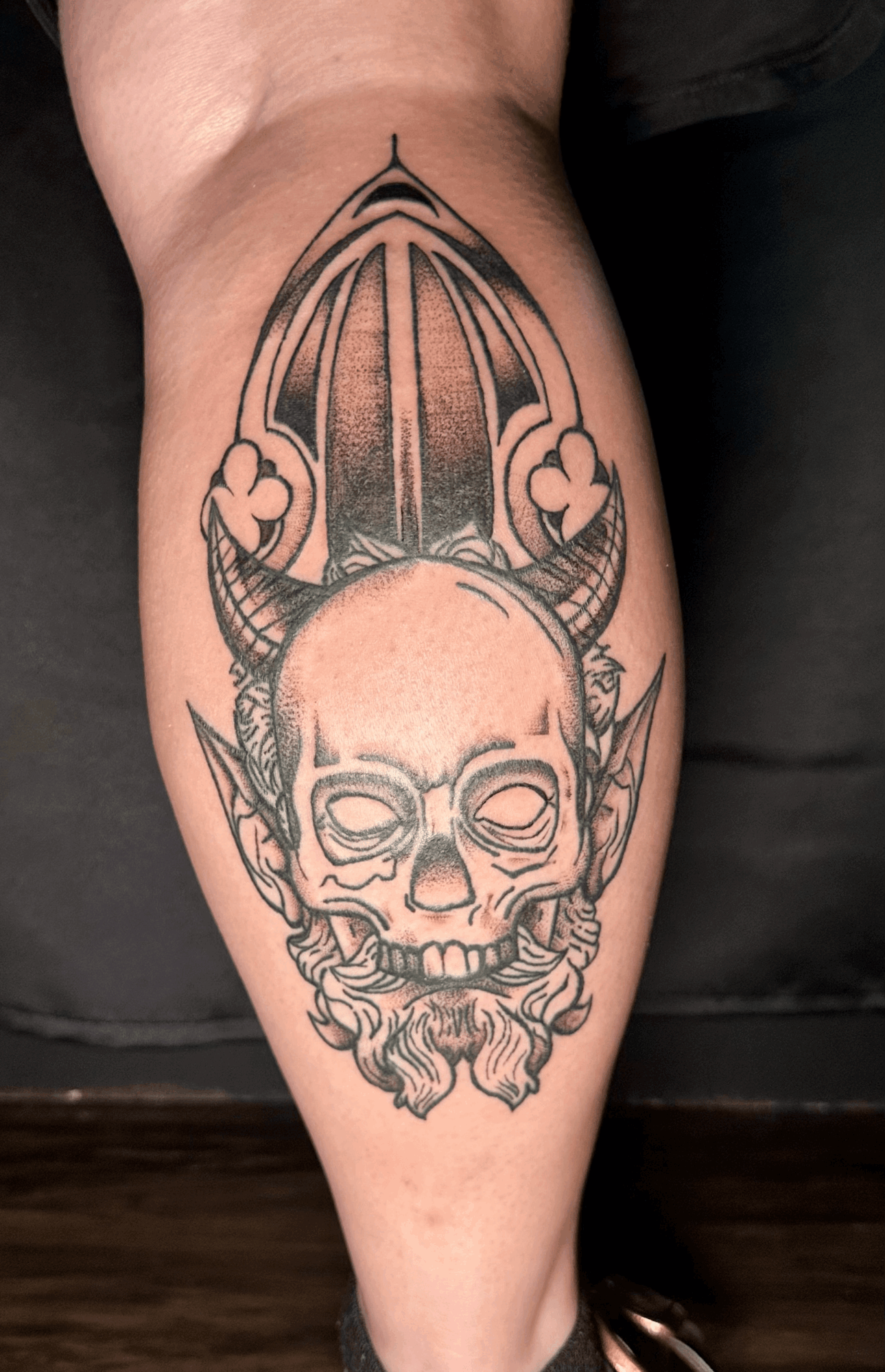 Tattoo of a skull with horns, pointed ears, and a beard, with a sword behind it, on a person's arm.