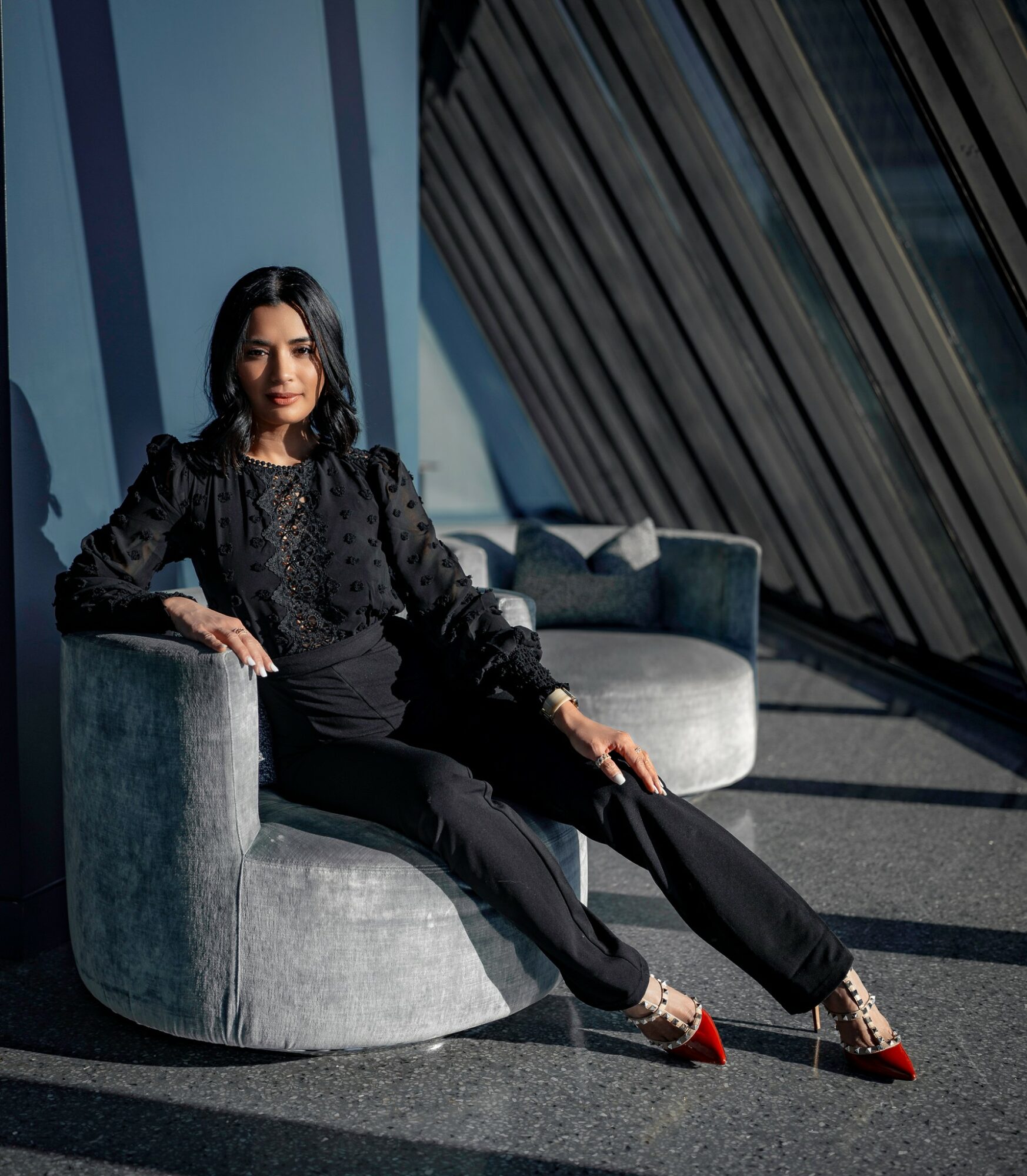 Woman sitting on a round gray sofa in a modern room with large windows and diagonal blinds, sunlight casting shadows.