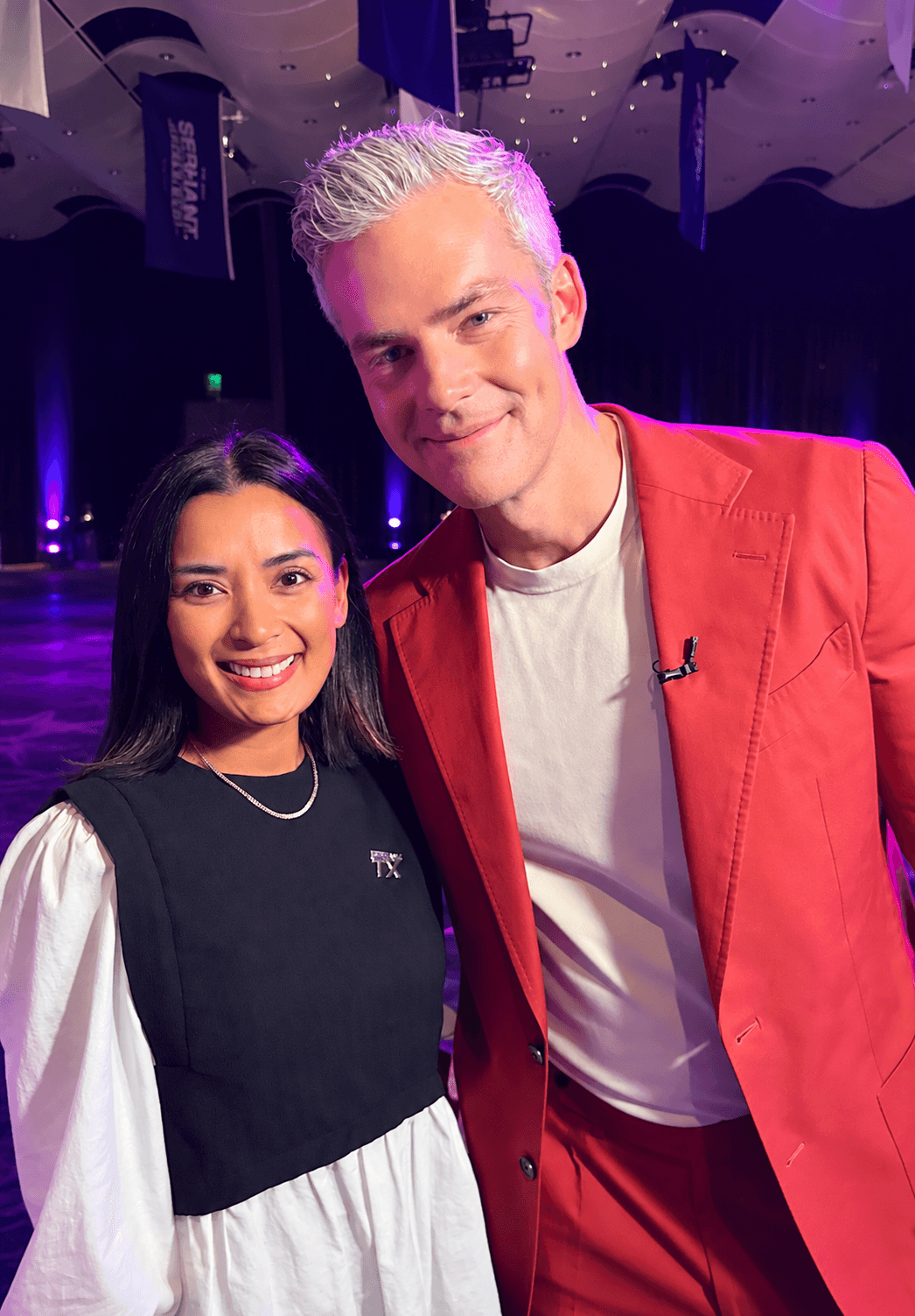 Two people smiling, one with dark hair and the other with light hair, in a dimly lit venue with purple lighting.