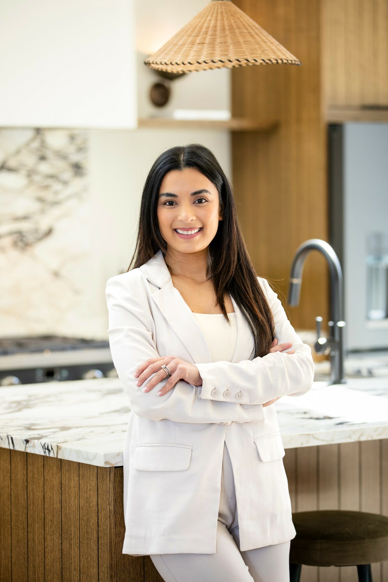 Smiling woman in white blazer standing in modern kitchen with marble countertop and wooden cabinets