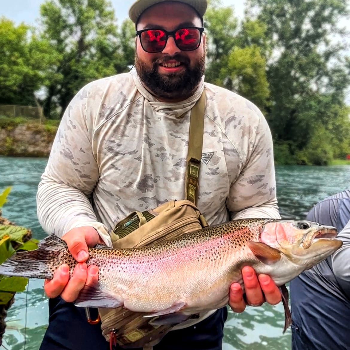 Man holding a large fish outdoors near water, wearing sunglasses, a cap, and a long-sleeve shirt, smiling.