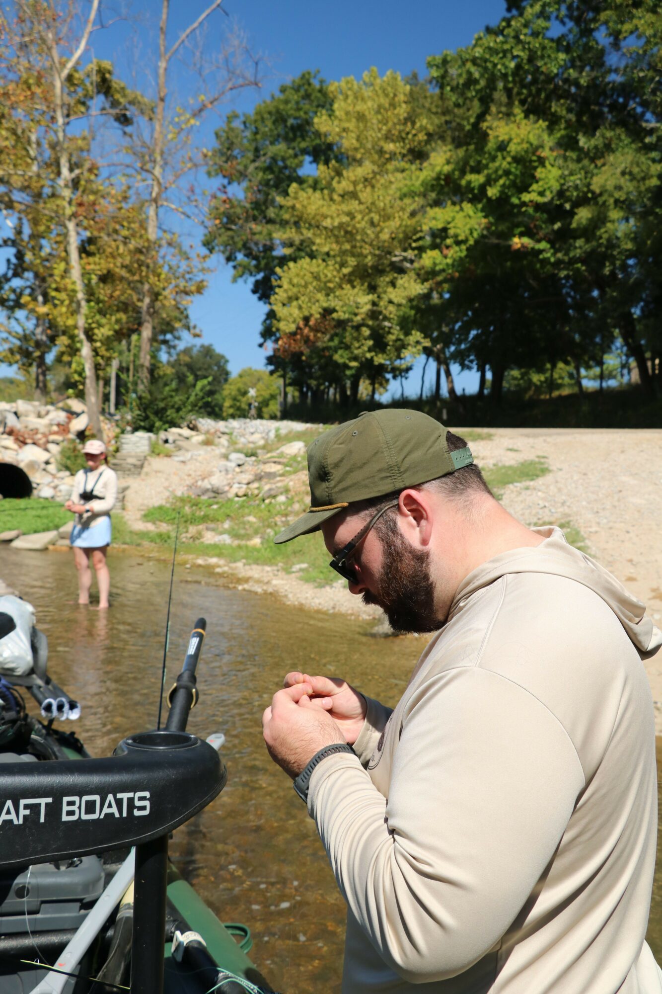 Man wearing a hat and sunglasses stands outdoors near trees and water, holding a drink, with people in the background.