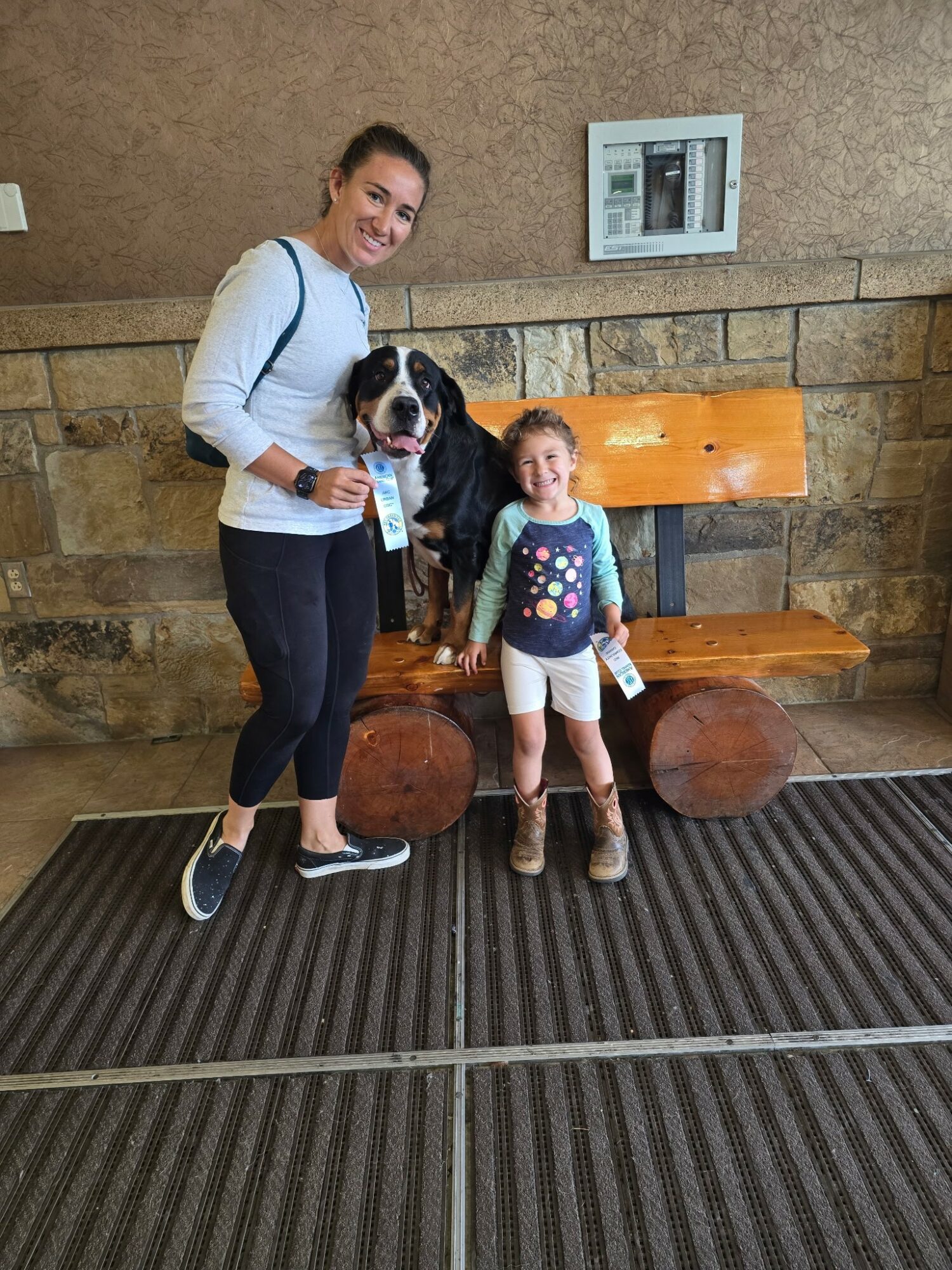 Woman and young girl with a dog sitting on a wooden bench against a stone wall, woman smiling, girl holding a paper, dog sitting between them.
