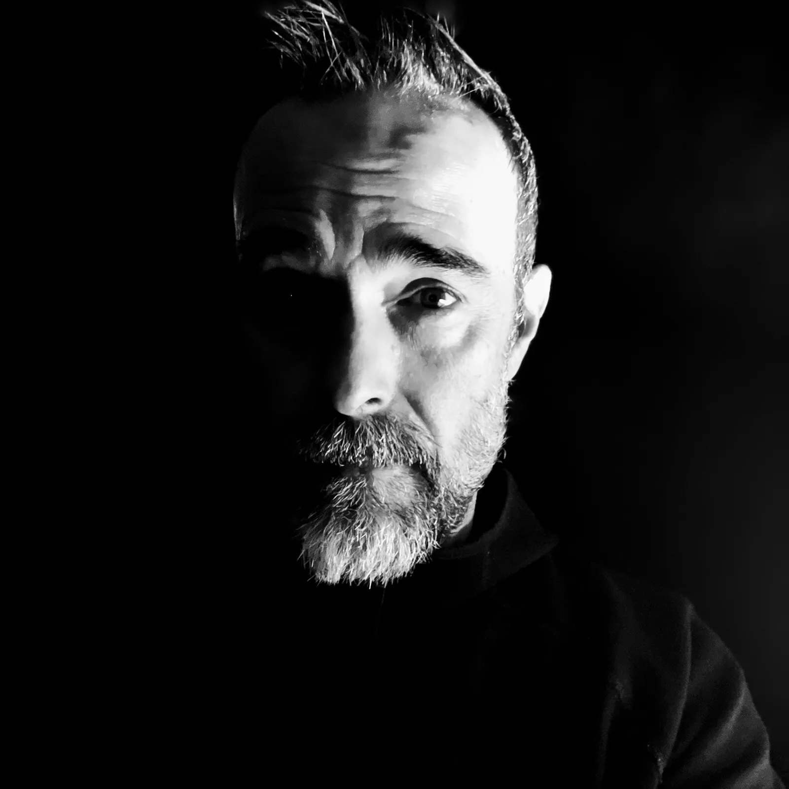 Portrait of a man with a beard, half-lit face, in black and white, looking directly at the camera.